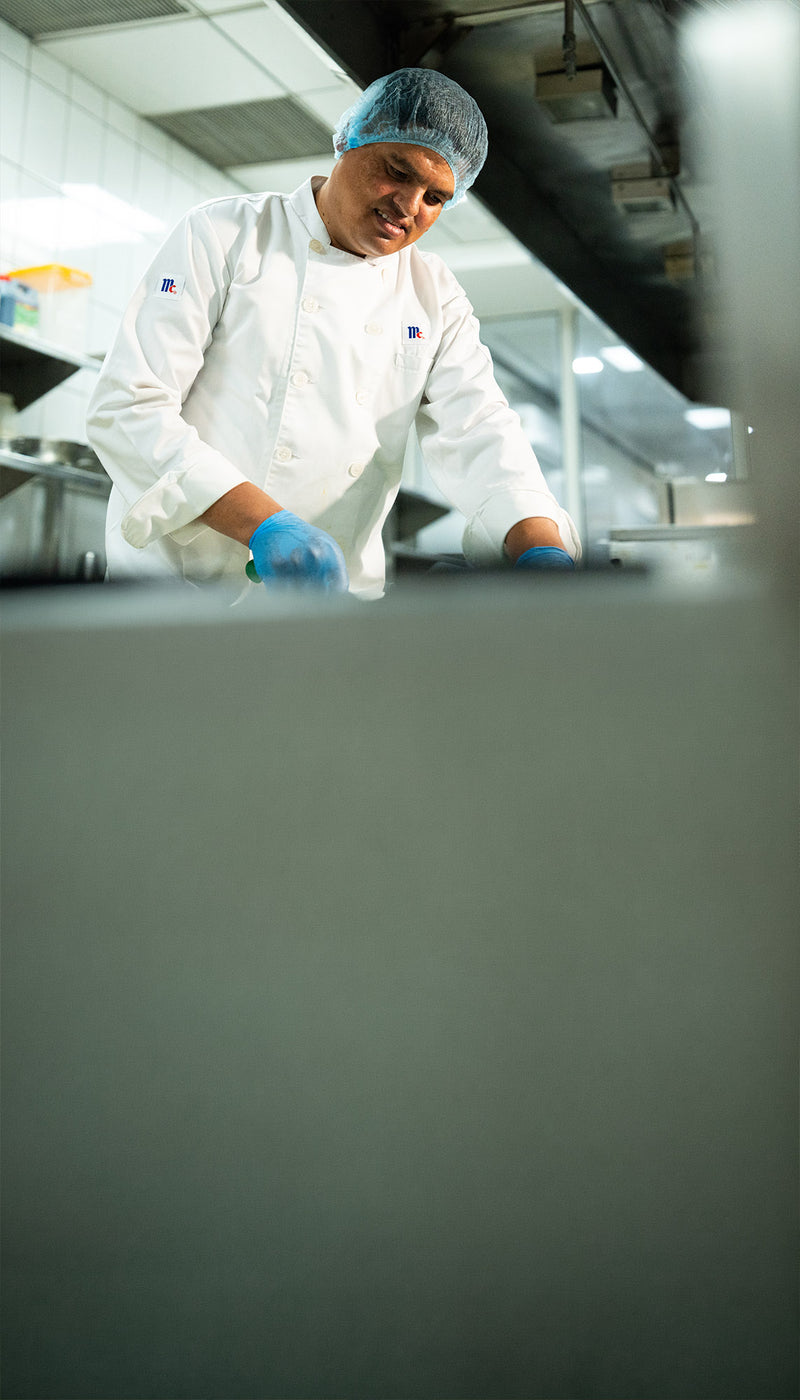 Chef in a white McCormick coat and hairnet preparing food in a commercial kitchen.