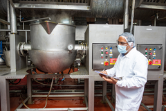 Person in a white lab coat and face mask using a tablet in a industrial kitchen setting.