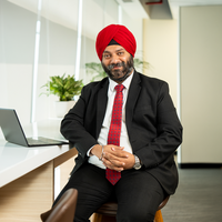 A man wearing a black suit, white dress shirt, red patterned tie, and a bright red turban sits on a stool in a modern office setting.