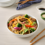 Bowl of noodles with vegetables and meat on a light surface with chopsticks and soy sauce.