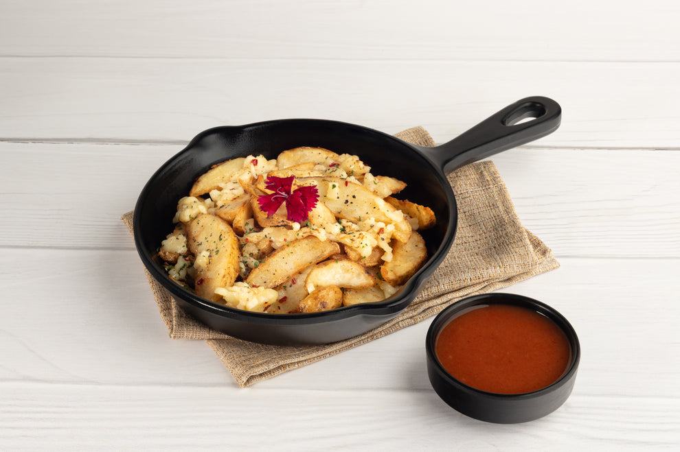 Fried potato in a black skillet with a side of sauce on a white wooden surface